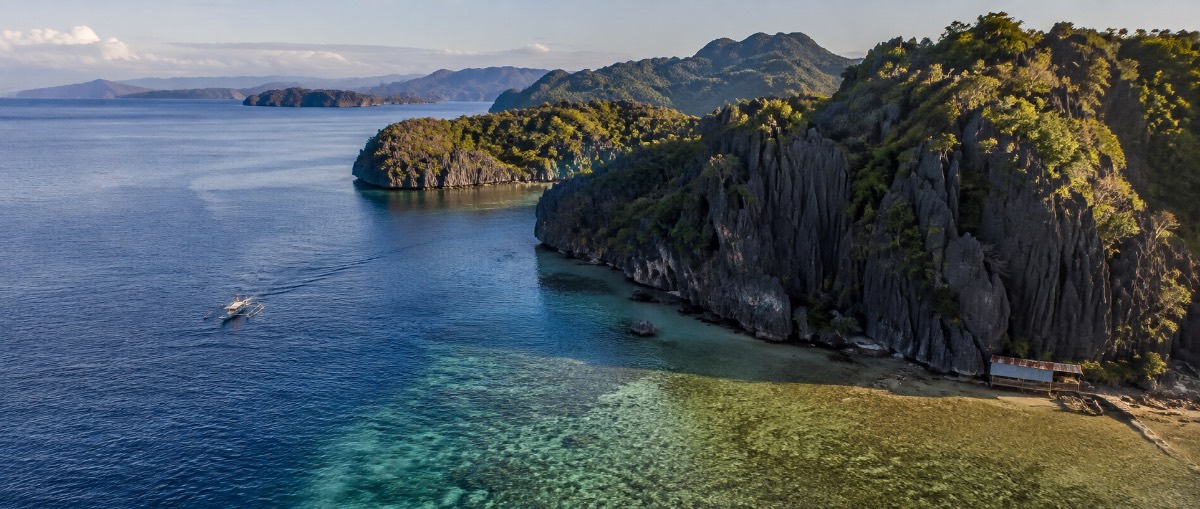 Limestone karst coastline in the Philippines at golden hour, with an outrigger bangka crossing a turquoise reef